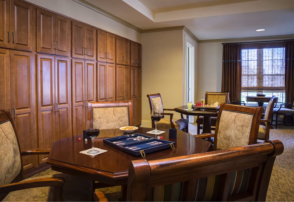Cozy interior common room with wooden tables and upholstered chairs, a backgammon set and wine glasses on the table, and large wooden cabinets along one wall.