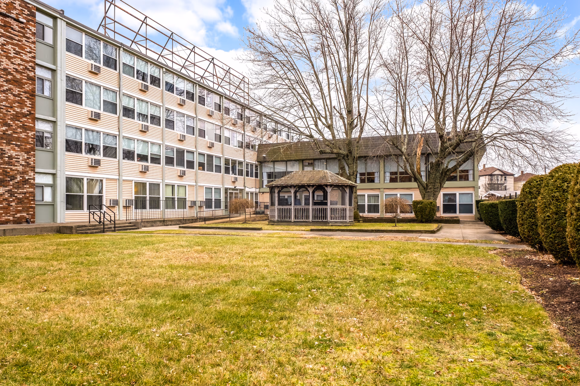 Outdoor view of River Falls Senior Living facility showing a multi-story building with numerous windows and air conditioning units. In front of the building is a grassy lawn with a wooden gazebo and leafless trees, under a partly cloudy sky.