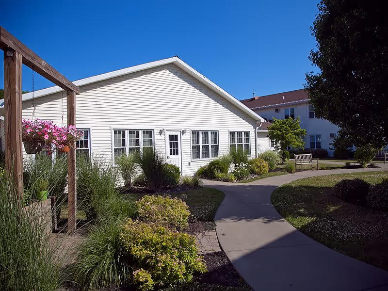 Outdoor view of a senior living facility with a white building, a paved walkway, green bushes, flowering plants, and a wooden pergola with hanging pink flowers under a clear blue sky.