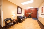 A waiting area with a black leather couch and a black chair separated by a small wooden table with a lamp. The walls are painted beige with a red accent wall at the far end. There is a decorative tree wall art on the red wall and framed pictures on the beige walls. The floor is light wood, and the ceiling has recessed lighting.