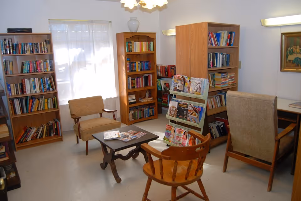A cozy reading room with several wooden bookshelves filled with books, a magazine rack with various magazines, and four chairs arranged around a small wooden table. The room has a window with sheer curtains allowing natural light to enter, and a painting on the wall.