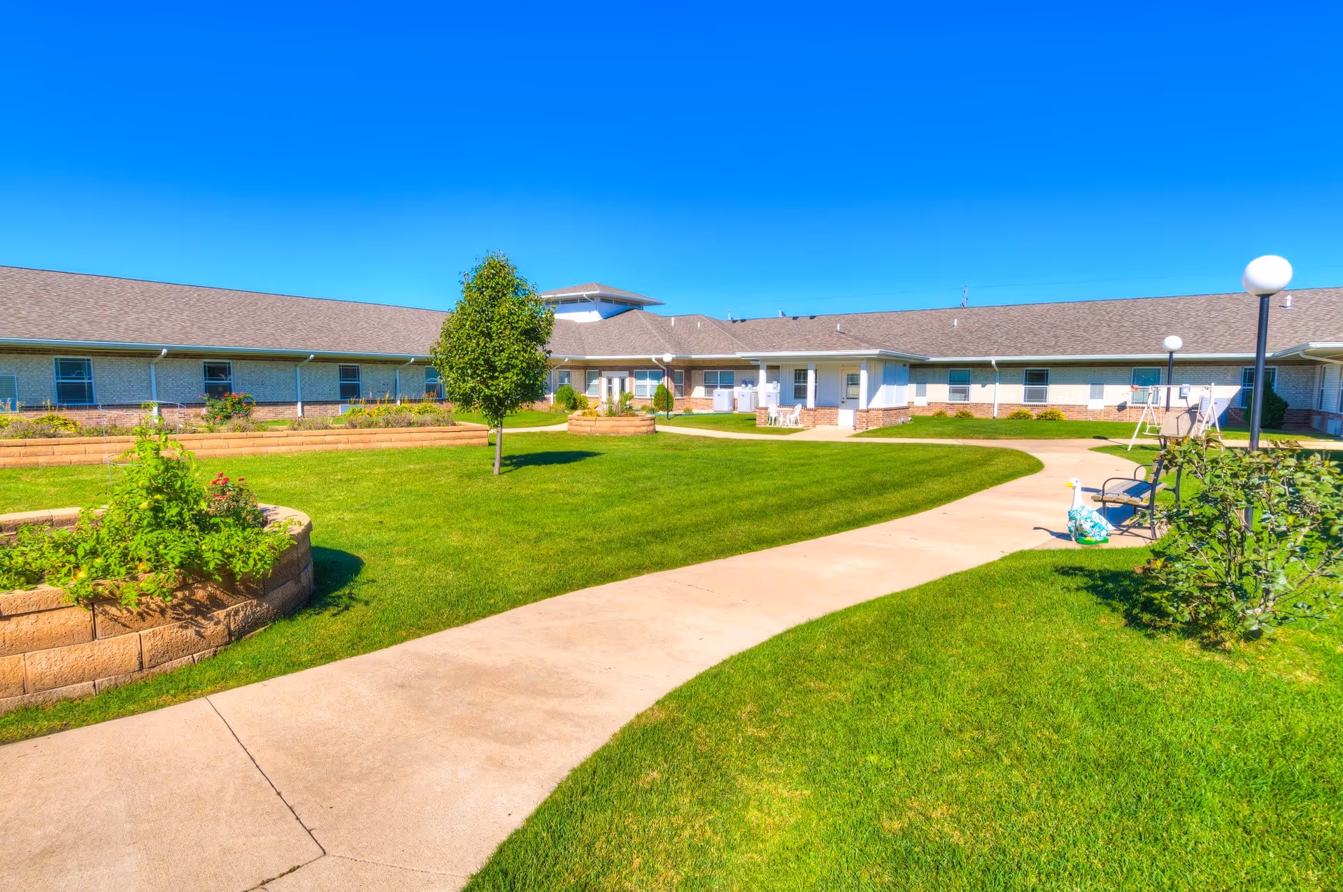 Outdoor courtyard area of a senior living facility with a curved concrete pathway, green grass, small trees, raised garden beds, benches, and a swing set. The building surrounds the courtyard with a clear blue sky overhead.