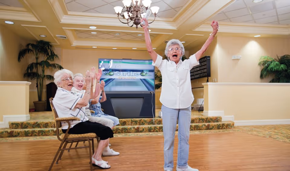 An elderly woman standing with her arms raised in celebration in a senior living facility game room, while three other elderly women sitting on chairs behind her clap and smile. The room has a carpeted floor, a large screen displaying a bowling game, and decorative plants.