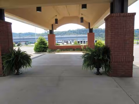 Senior living facility entrance under a covered walkway with brick columns, potted plants, and distant parking.
