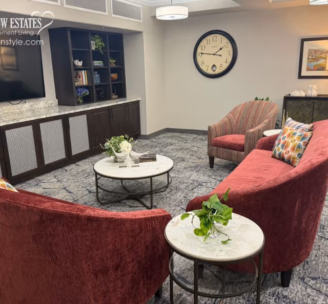 A cozy living room area with two curved red sofas and a striped armchair arranged around two round marble-top coffee tables with green plants. A large wall clock hangs on a beige wall above a dark wood cabinet with shelves containing books and decorative items. The floor is carpeted with a patterned design, and a flat-screen TV is mounted on the wall to the left.