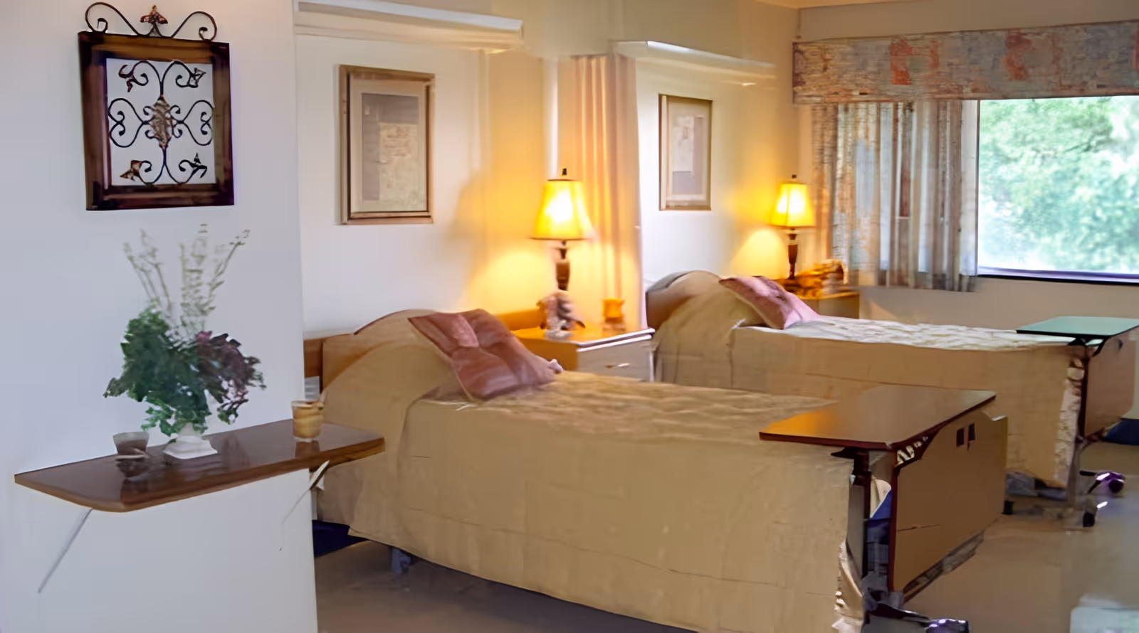 A nursing facility room with two hospital-style beds covered in beige bedspreads, each with a pink pillow. There are two bedside tables with lamps emitting warm light. A window with patterned curtains lets in natural light. A small table with a vase of flowers is visible in the foreground, and framed artwork hangs on the walls.