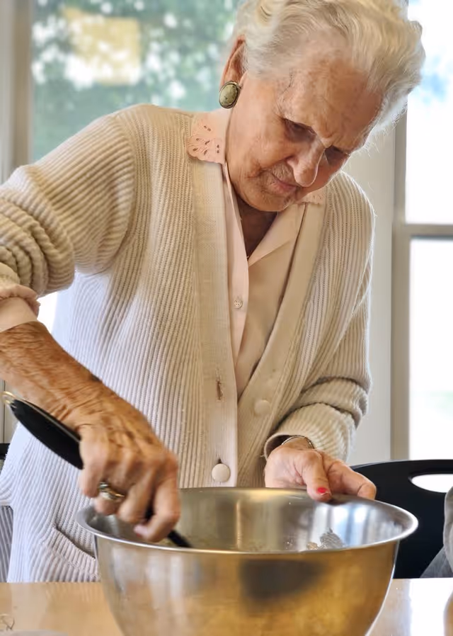 An elderly woman wearing a light-colored cardigan and earrings is stirring the contents of a large metal mixing bowl on a table in a well-lit room with windows in the background.