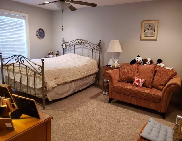 A cozy bedroom featuring a metal-framed bed with a beige quilt, a brown upholstered loveseat with stuffed animals on it, a wooden nightstand with a lamp, and framed pictures on the walls. The room has beige carpeting and a ceiling fan with a light fixture.