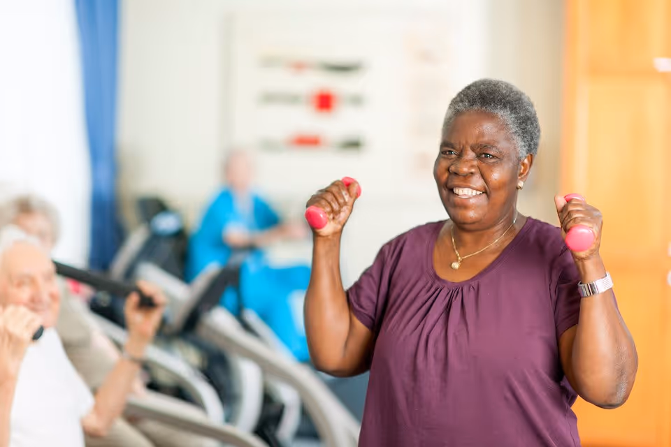 An elderly woman smiling and holding small pink dumbbells while exercising indoors, with other seniors using exercise equipment in the background.