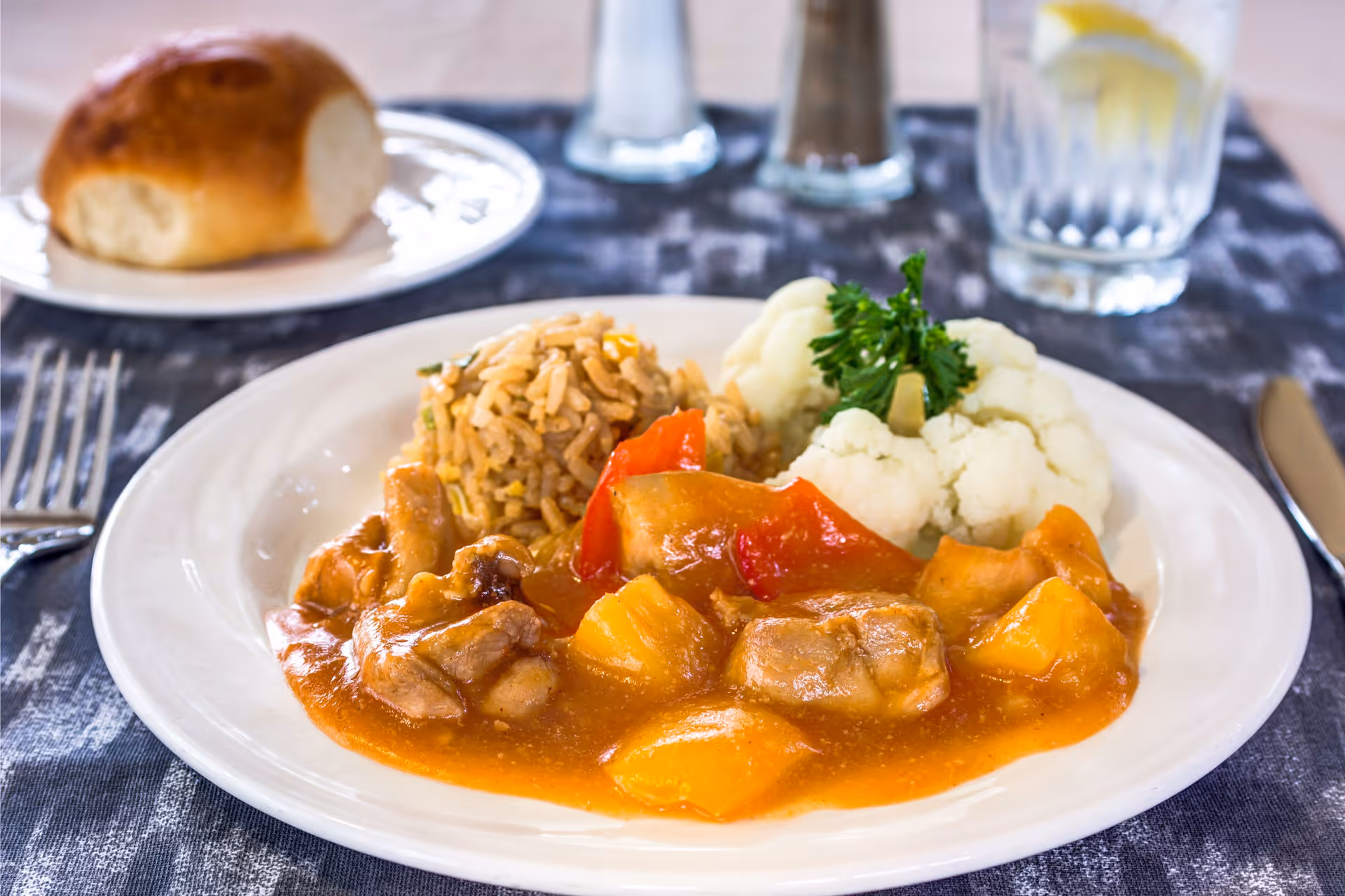 A plate of food featuring a serving of rice, cauliflower garnished with parsley, and a stew with chunks of meat, pineapple, and red bell peppers in a reddish sauce. In the background, there is a bread roll on a small plate, a glass of water with lemon, and salt and pepper shakers on a dark patterned tablecloth.