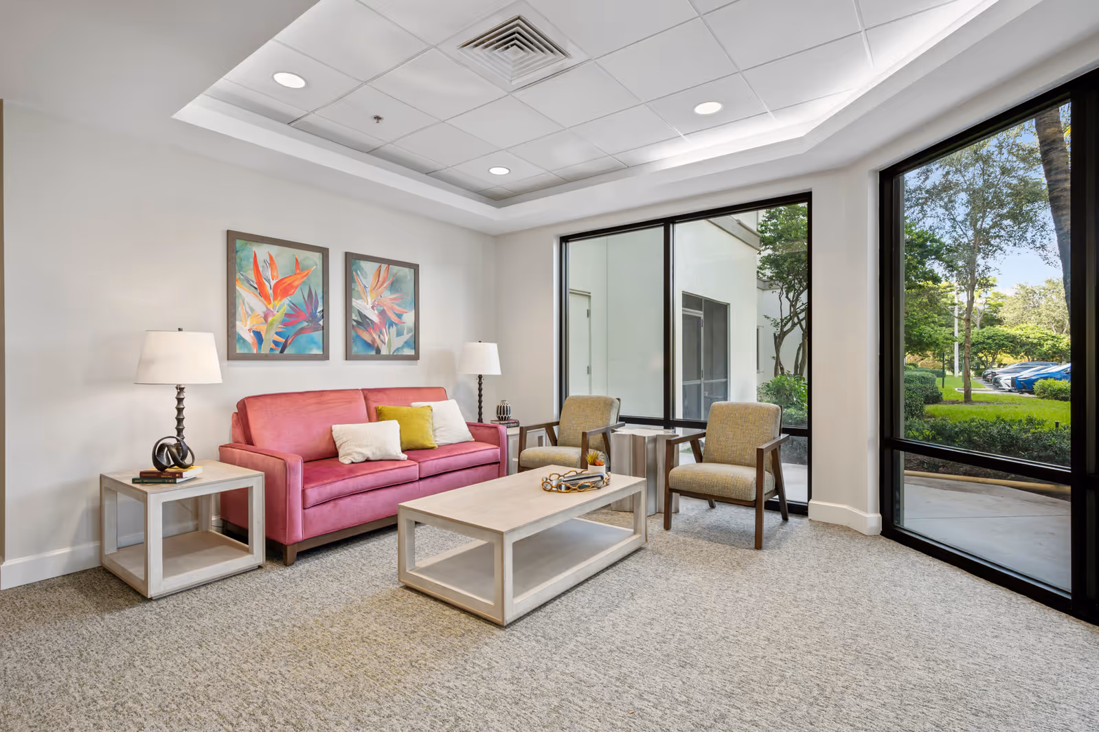 A bright and modern living room with a pink sofa adorned with three pillows, two beige armchairs, a light wood coffee table, and two matching side tables with lamps. The room features large windows showing a green outdoor area with trees and parked cars. Two colorful floral paintings hang on the white wall above the sofa.