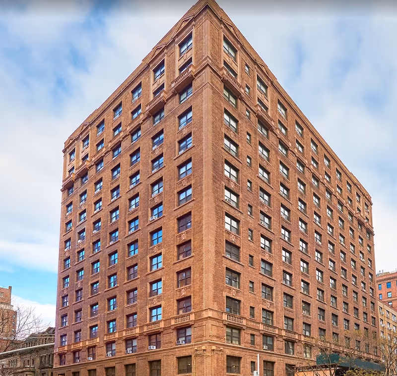 Tall multi-story brick building with many windows under a partly cloudy sky, viewed from a corner angle.