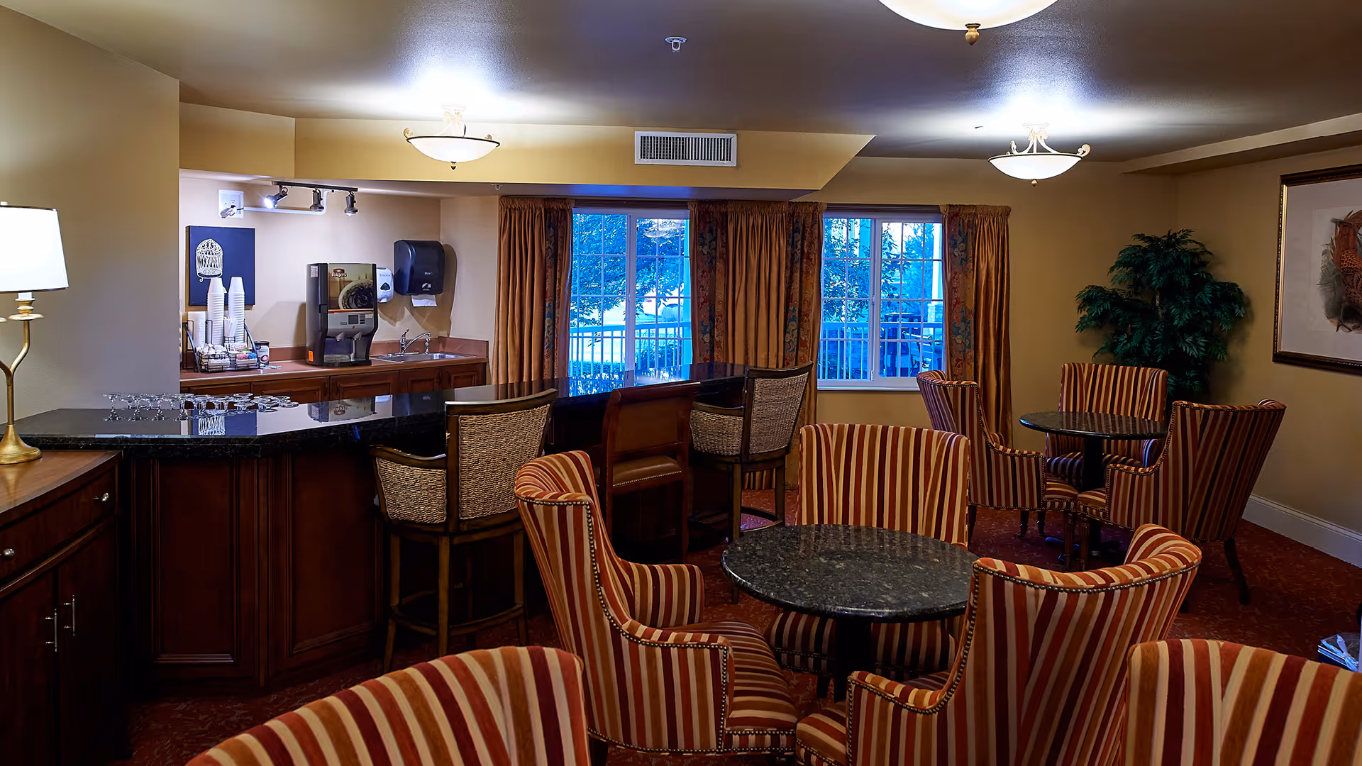 Cozy common room with striped armchairs around small round tables, a service counter with bar stools, and windows with curtains.