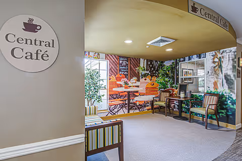 Interior view of a senior living community café area named Central Café, featuring a cozy seating arrangement with striped chairs and a mural depicting an outdoor café scene with tables, chairs, and greenery.