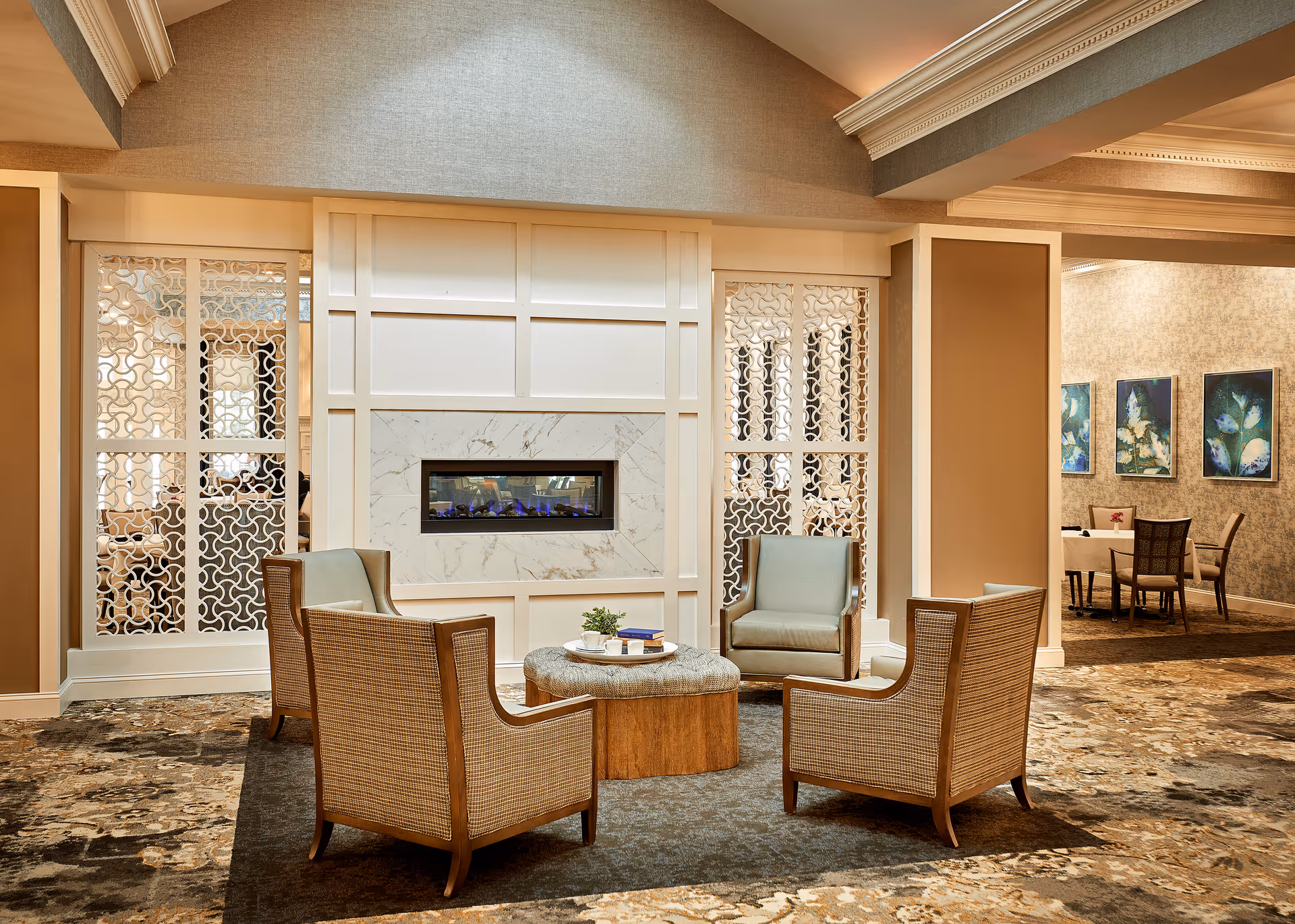 A cozy seating area in a senior living facility featuring four cushioned armchairs arranged around a round ottoman with a small plant and books on top. The seating area is set on a patterned carpet in front of a modern electric fireplace with a white paneled wall and decorative lattice panels on either side. In the background, there is a dining area with tables, chairs, and framed floral artwork on the wall.