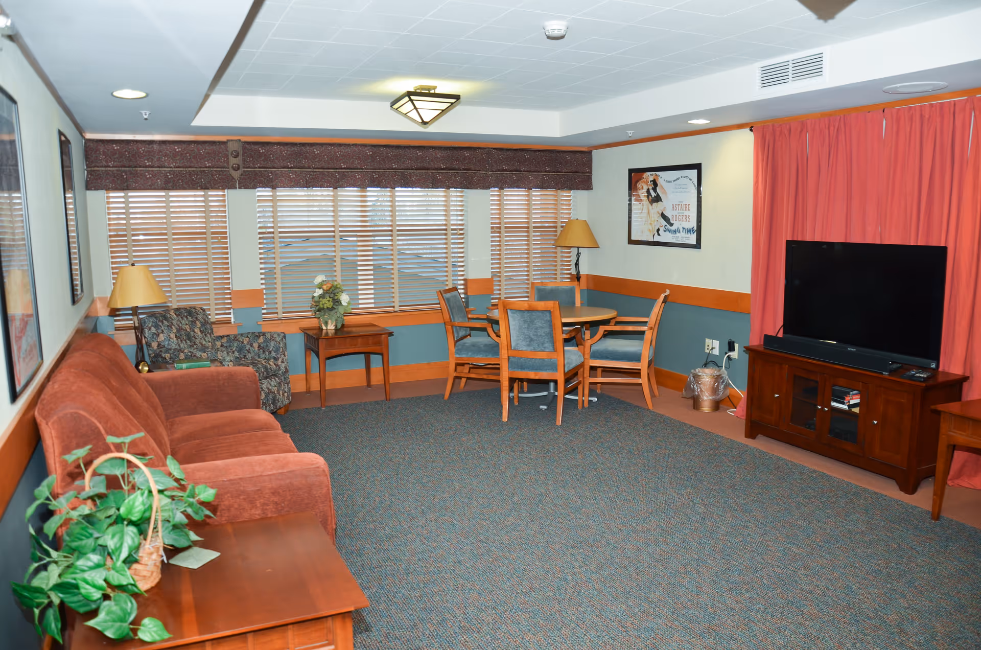 A cozy living room with a rust-colored sofa, a patterned armchair, a wooden side table with a lamp, and a small round dining table with four chairs. The room has large windows with wooden blinds, a TV on a wooden stand, red curtains, and framed artwork on the walls.