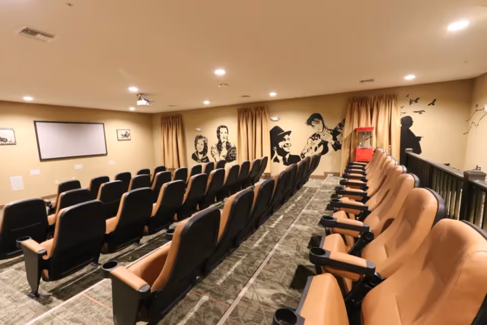 A small theater room with two rows of tan and black cushioned seats facing a blank projector screen on a beige wall. The walls are decorated with black silhouette murals of classic movie characters and scenes. There is a red popcorn machine in the back corner of the room, and the floor is carpeted with a patterned design.
