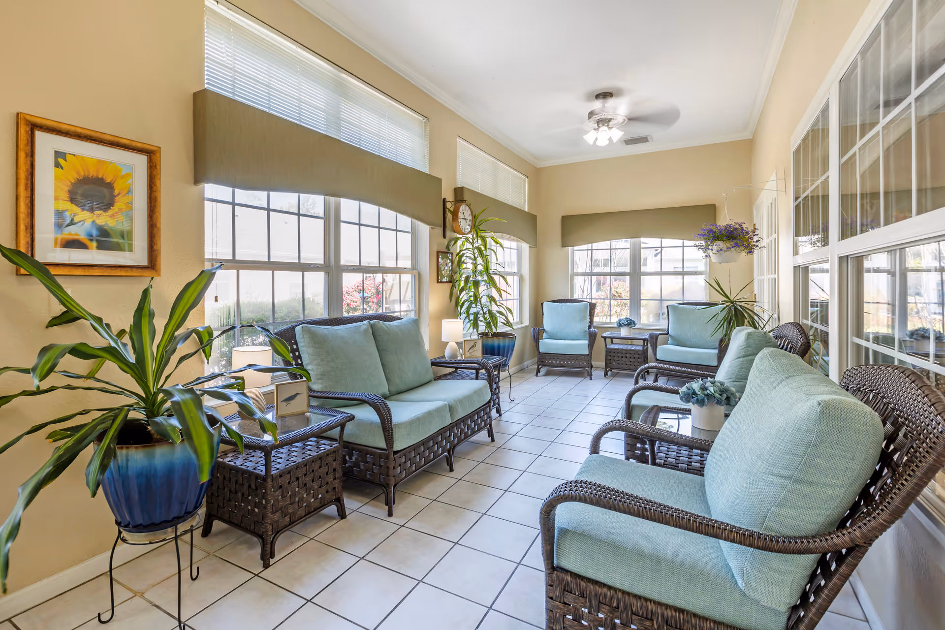 A bright sunroom with large windows on two sides, furnished with wicker chairs and loveseats with light blue cushions. There are several potted plants, a small side table with a lamp, and a framed sunflower picture on the wall. The floor is tiled and the ceiling has a fan with lights.