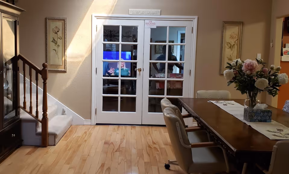 Interior view of a dining area with a wooden table and cushioned chairs on wheels. A vase with flowers and a tissue box are on the table. To the left, there is a staircase with wooden railings and a glass cabinet. Two framed floral paintings hang on the walls. In the background, white French doors with glass panes lead to another room where a television is visible.