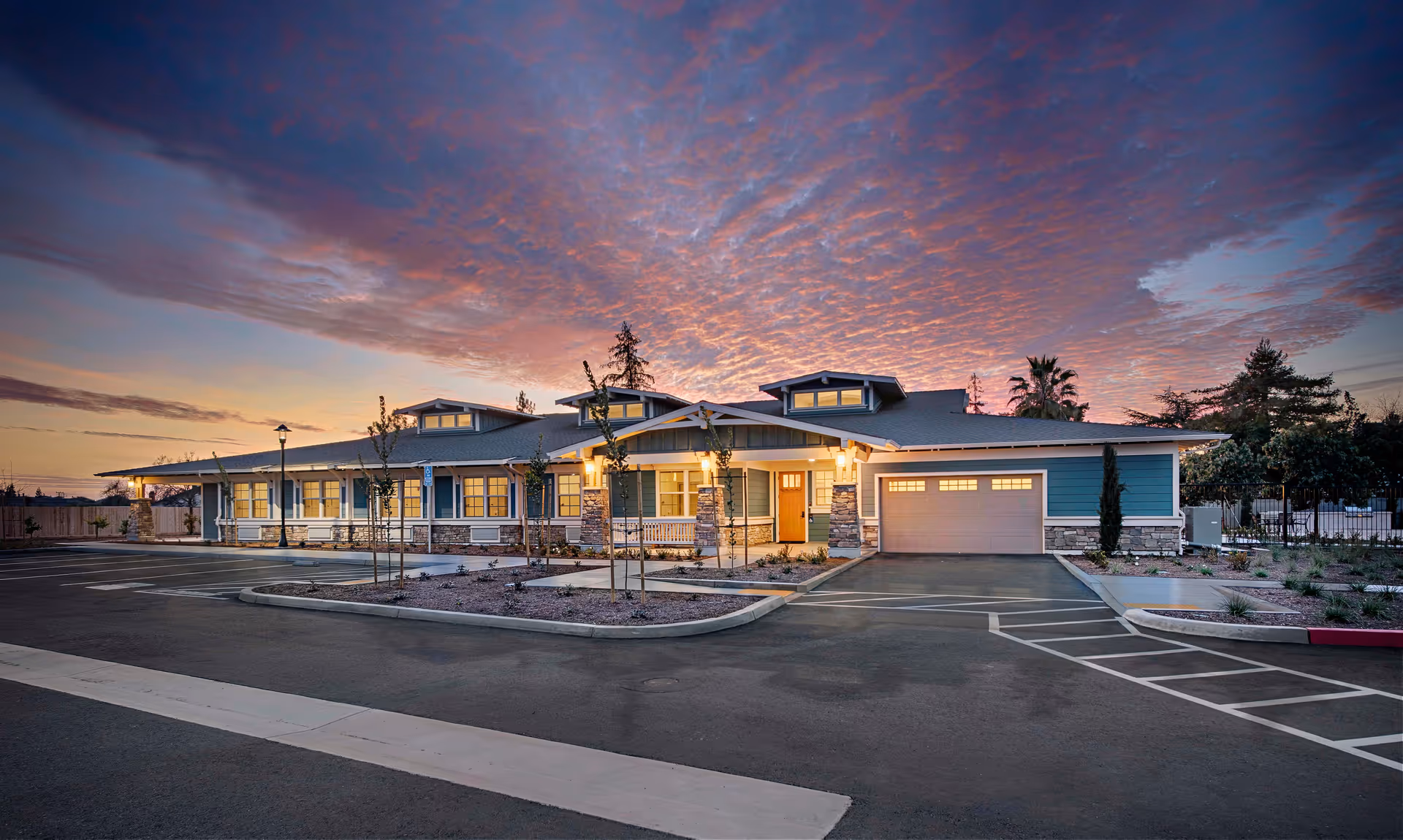 Exterior view of Magnolia Crossing Assisted Living facility at dusk, showing a single-story building with blue siding, stone accents, and a well-lit entrance. The sky is colorful with a dramatic sunset and there is a parking area in front with marked spaces and young trees planted along the walkway.
