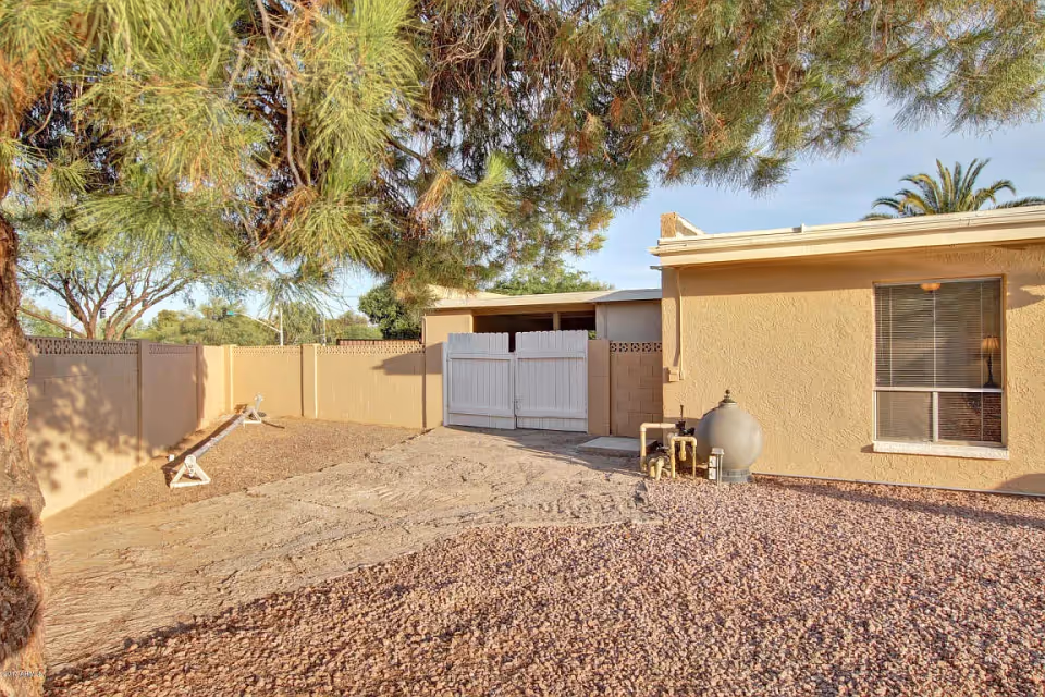 Outdoor area of a residential facility with a gravel ground, a large tree on the left, a beige stucco building with a window on the right, and a white double gate in the center attached to a beige block wall fence.