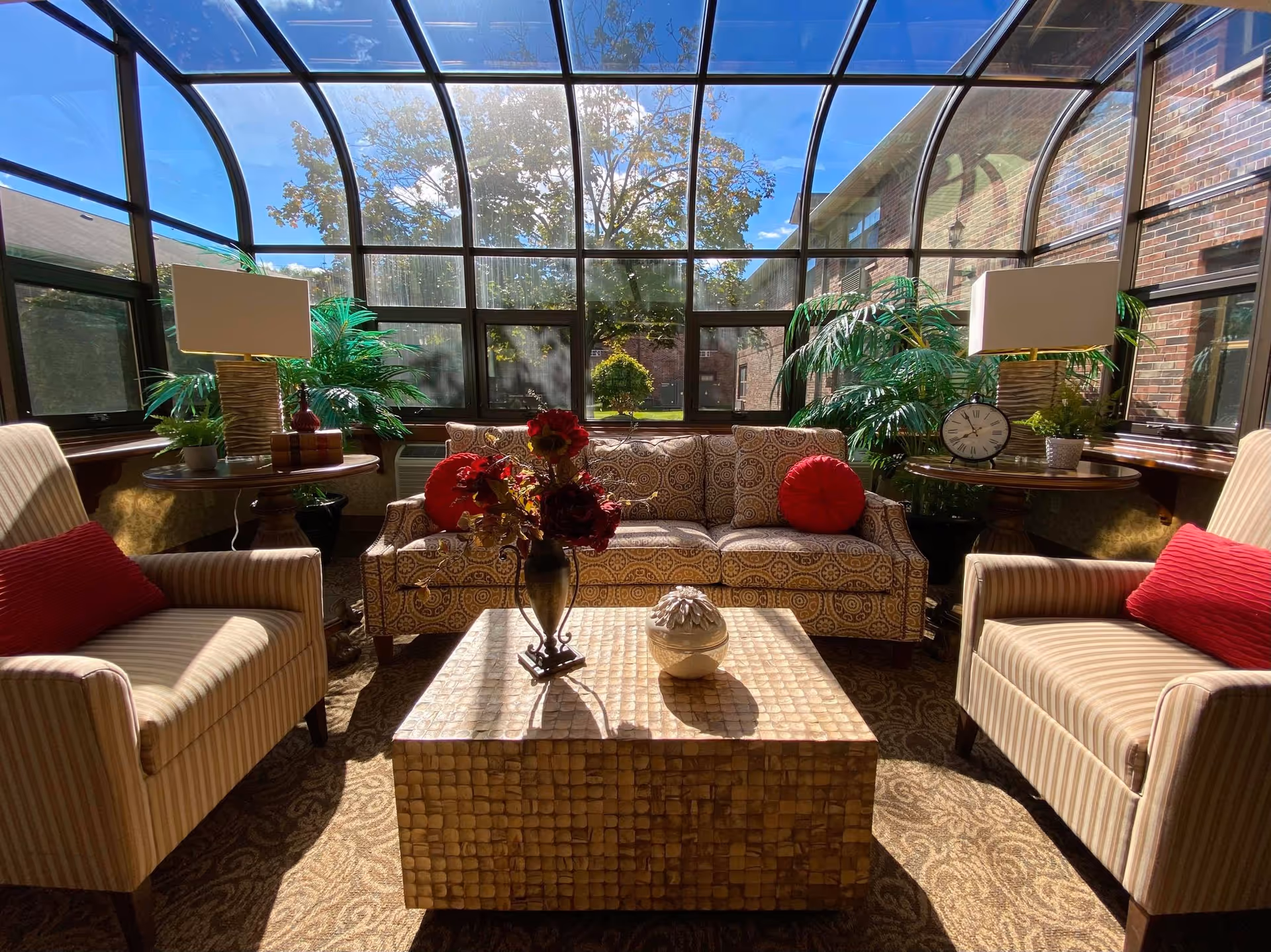 A sunlit indoor seating area with a glass ceiling and walls showing an outdoor view. The room features a patterned sofa with red round pillows, two striped armchairs with red cushions, two wooden side tables with lamps and plants, and a square coffee table with decorative items. Green plants are placed near the windows, and the floor is carpeted with a patterned design.