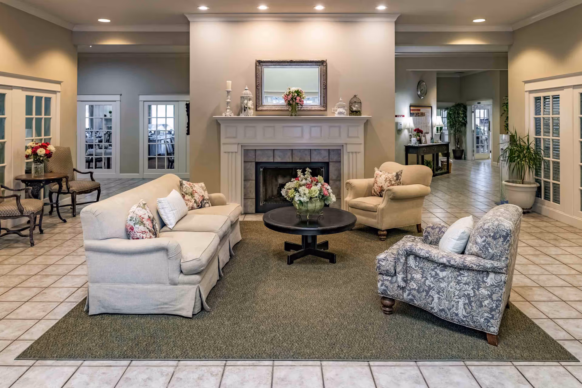 A cozy living room area in a senior living facility featuring a beige sofa, two armchairs with floral and patterned cushions, and a round black coffee table with a floral centerpiece. The room has a fireplace with decorative items and a mirror above it. The floor is tiled with a large area rug under the seating arrangement. There are additional chairs and tables along the sides, and the space is well-lit with recessed ceiling lights.