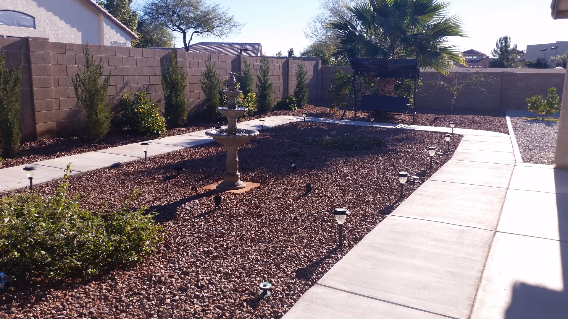 Outdoor garden area with a stone pathway, small shrubs, a multi-tiered water fountain in the center, and a black metal bench swing under a palm tree, all enclosed by a tall brick wall.