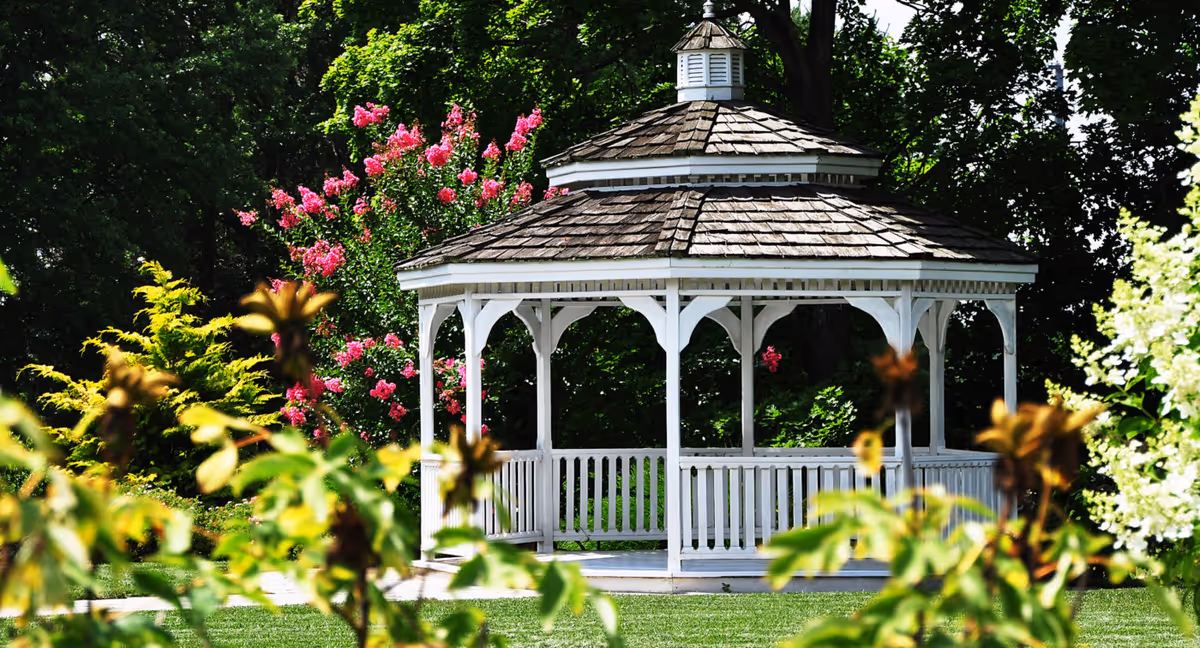 A white wooden gazebo with a shingled roof situated in a lush garden surrounded by green trees and vibrant pink and yellow flowers.
