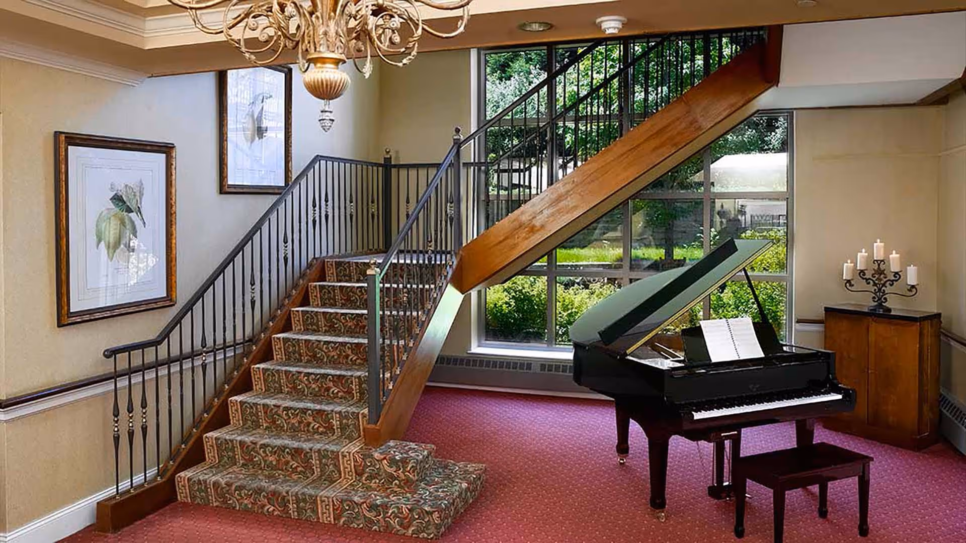Interior view of a senior living facility featuring a carpeted staircase with ornate railing, a black grand piano with an open sheet music book, a wooden cabinet with a decorative candle holder, framed botanical artwork on the wall, a chandelier, and large windows showing greenery outside.