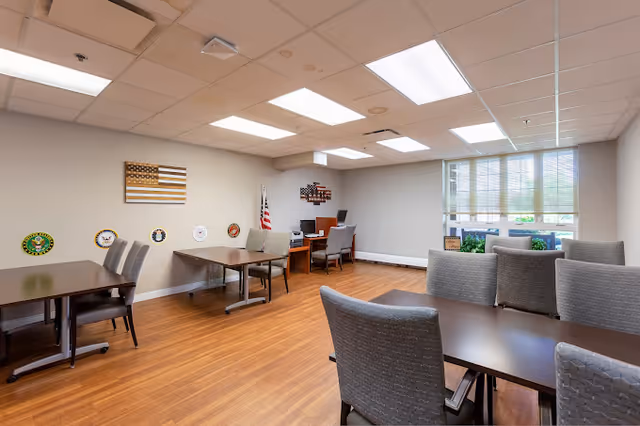Bright community room with multiple tables and upholstered chairs, patriotic wall decor and an American flag by a window.