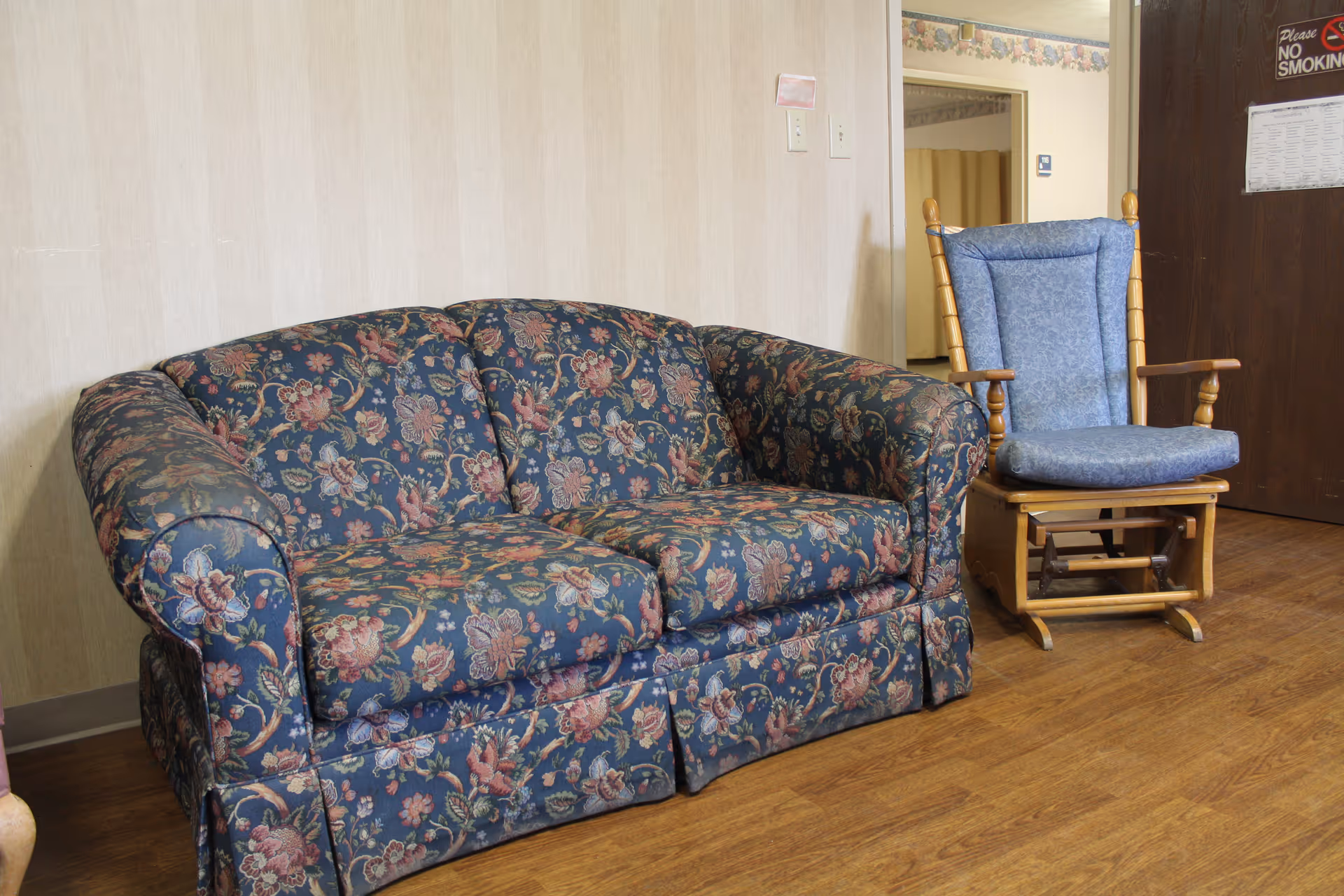 A floral patterned blue couch and a wooden rocking chair with blue cushions are placed on a wooden floor against a beige wall. A dark brown door with a 'Please No Smoking' sign is visible in the background.