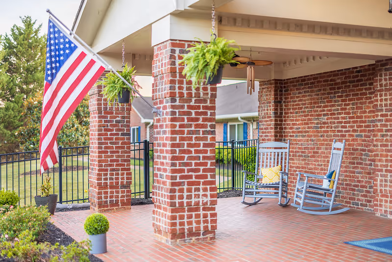 Covered brick front porch with an American flag, hanging ferns and two rocking chairs.