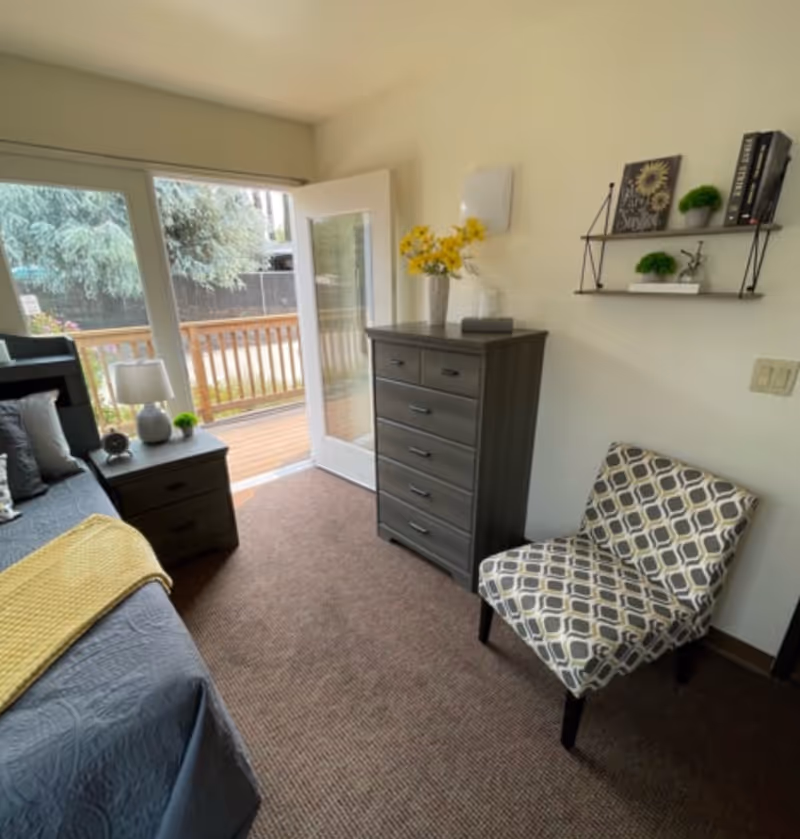 Small bedroom with a bed, nightstand, tall dresser, patterned chair and a glass door opening to an outdoor deck.