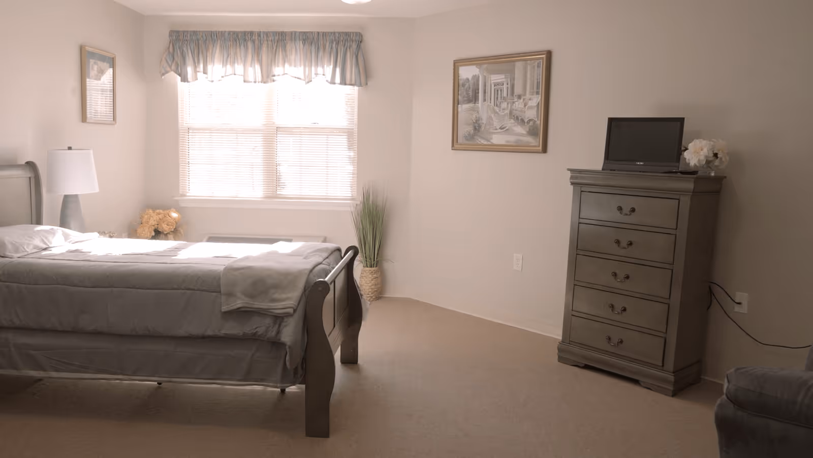 A bedroom with a bed covered in gray bedding positioned near a window with blinds and a blue valance. There is a nightstand with a lamp and flowers to the left of the bed. On the right side of the room, there is a tall gray dresser with a small TV and a flower arrangement on top. The walls are light-colored and decorated with framed artwork. The floor is carpeted.