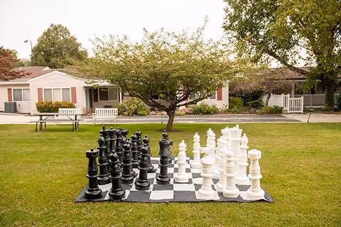 A large outdoor chess set with oversized black and white chess pieces arranged on a checkered board on a grassy lawn. In the background, there are trees, benches, and single-story buildings with white walls and red shutters.