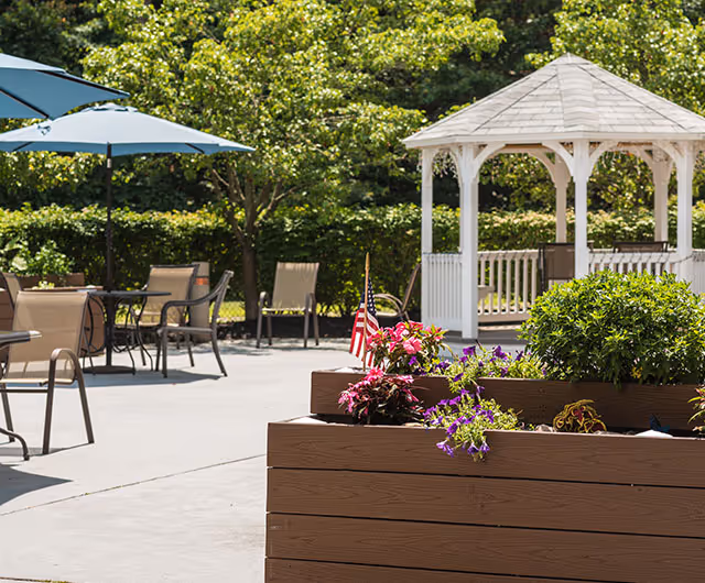 Outdoor patio area with tables, chairs, and umbrellas, surrounded by greenery and trees. A white gazebo is visible in the background, and a planter box with colorful flowers and a small American flag is in the foreground.