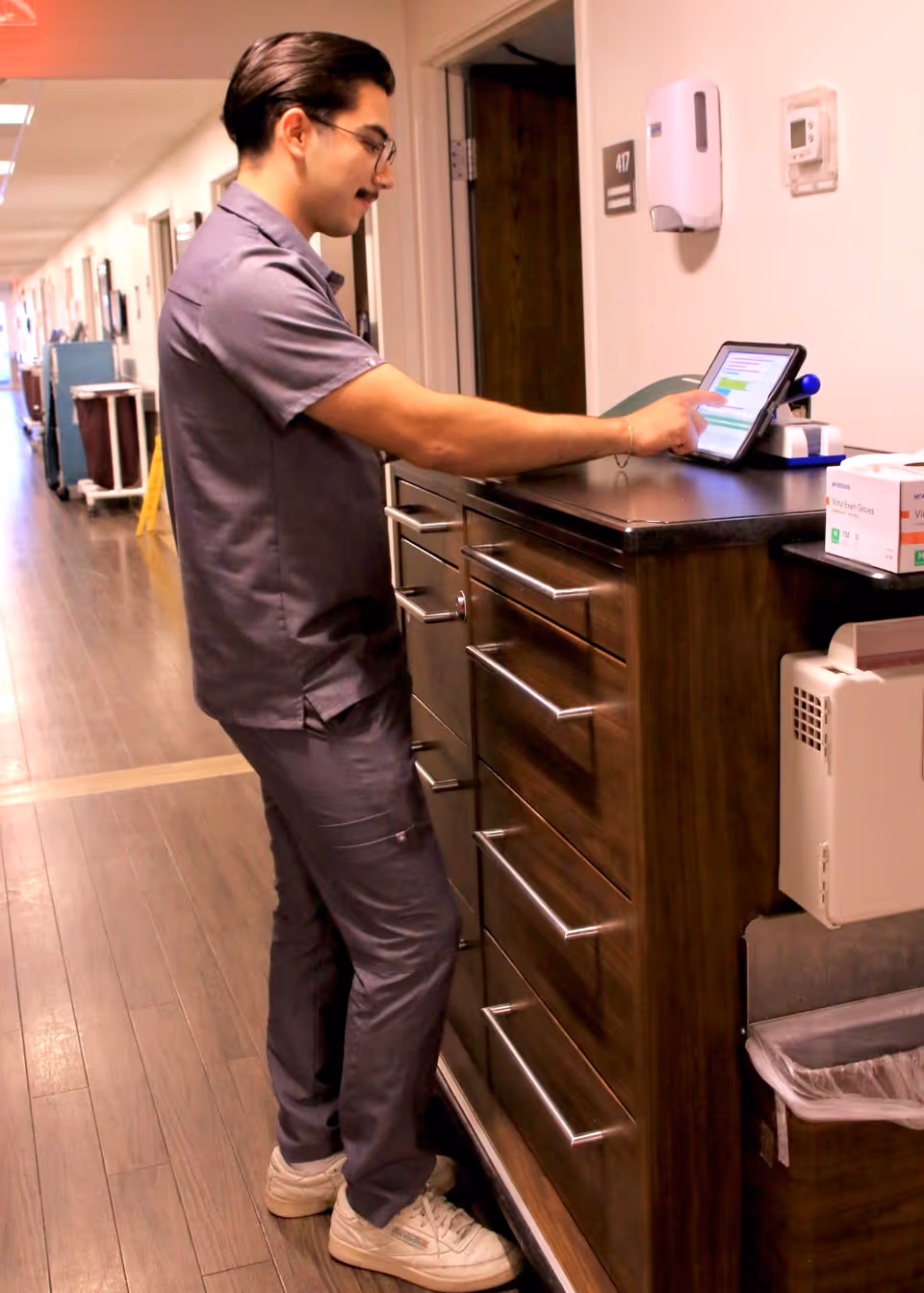 A healthcare worker in gray scrubs stands in a hallway, interacting with a tablet device placed on a wooden cabinet with multiple drawers. The hallway has wooden flooring and several doors along the side, with medical equipment and supplies visible in the background.