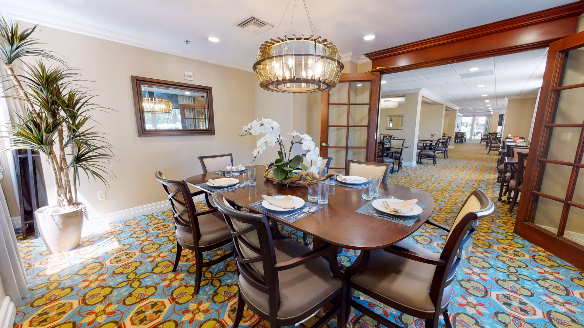 A dining area in a senior living facility featuring a wooden dining table set for six with plates, napkins, and glasses. The room has a colorful patterned carpet, a large potted plant, a wall mirror, and a modern chandelier hanging above the table. In the background, there is a hallway with more tables and chairs, separated by wooden framed glass doors.