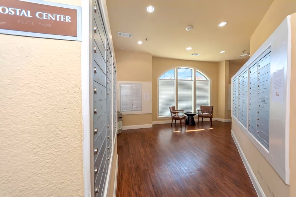 Interior view of a postal center room with multiple mailboxes mounted on both walls. The room has wooden flooring, beige walls, and a large arched window with blinds. Two wooden chairs and a small round table are placed near the window.