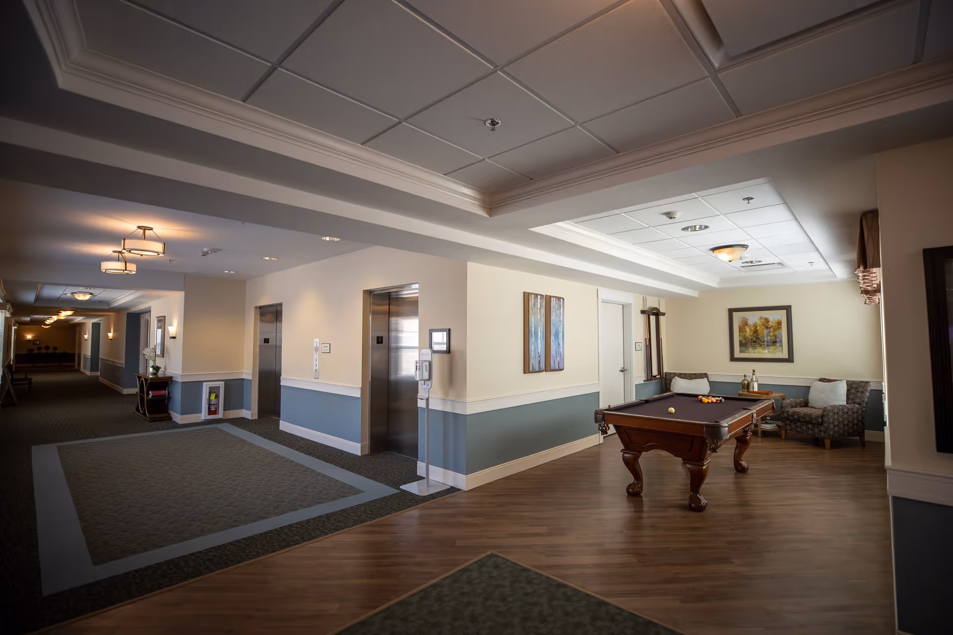 Interior view of a senior living facility hallway with two elevators on the left side and a small recreational area on the right featuring a pool table, two armchairs, a small table with bottles, and framed artwork on the walls.