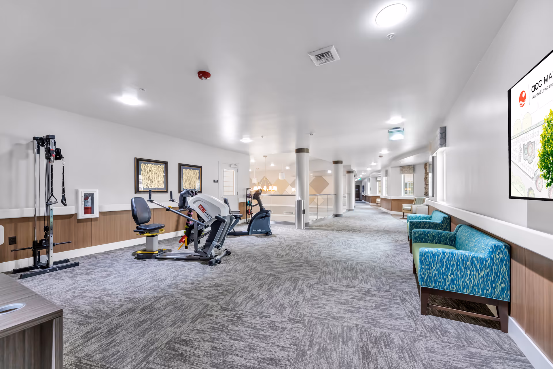 A spacious, well-lit hallway in a senior living community featuring exercise equipment on the left side, including a seated leg press and a stationary bike. On the right side, there are blue patterned armchairs and a large digital screen mounted on the wall displaying the facility's name and a map. The hallway has carpeted flooring, white walls, and ceiling lights, with windows along the right side letting in natural light.