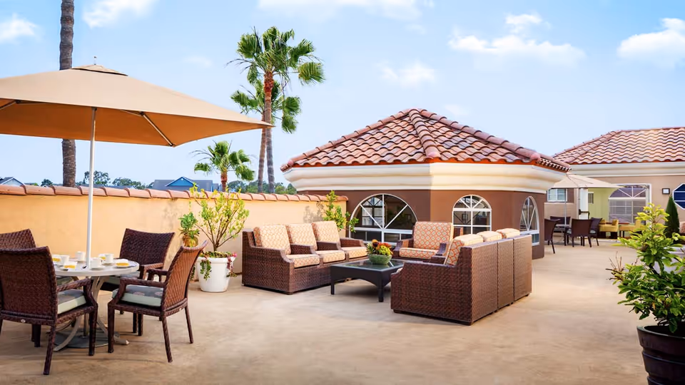 Outdoor patio area with wicker furniture including cushioned sofas and chairs, a table with an umbrella, potted plants, and a building with a tiled roof and arched windows under a blue sky with some clouds.