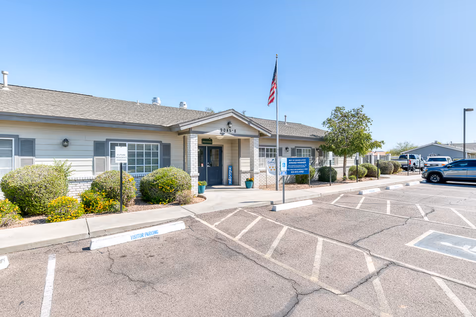 Single-story senior living facility entrance with an American flag, shrubs, and visitor parking in front.