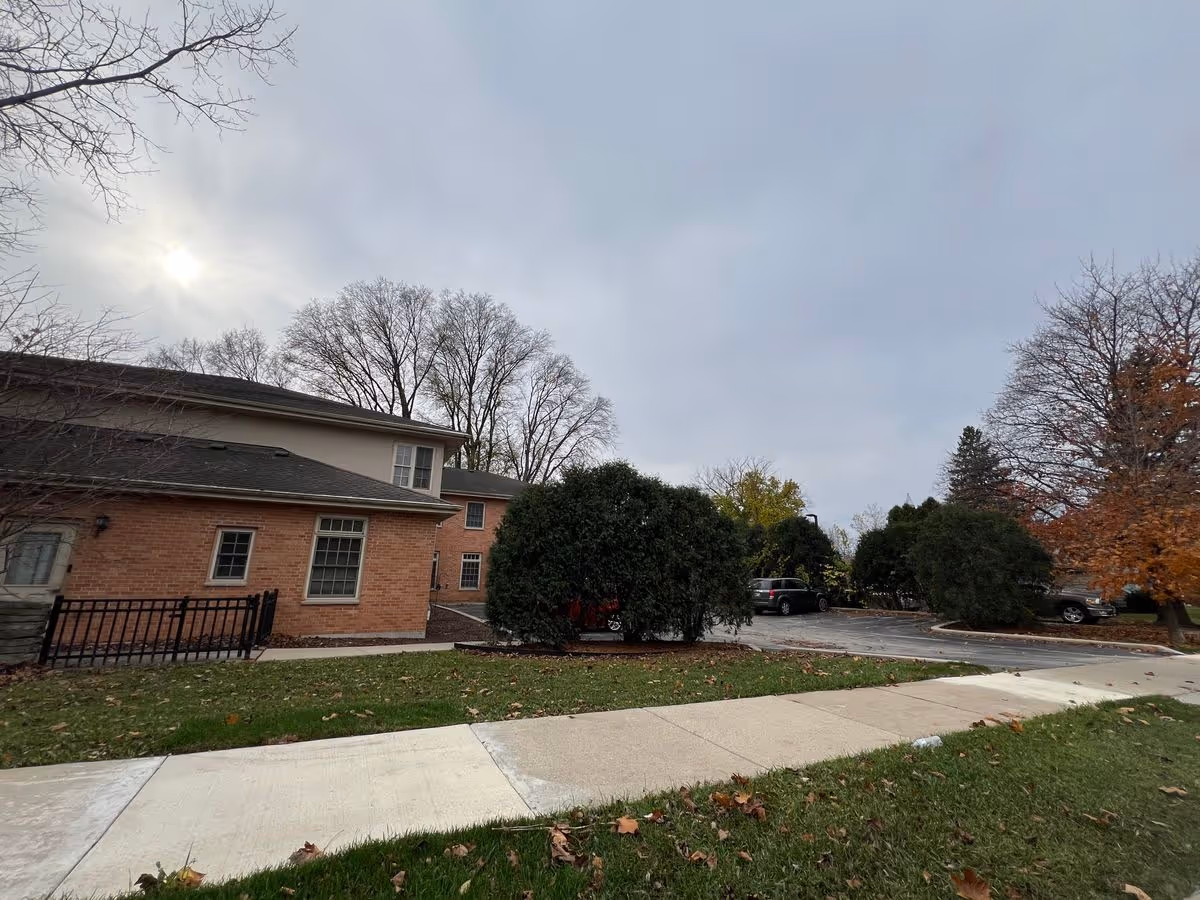 Front exterior of a brick senior living building with a sidewalk, parking area, and trees under a cloudy sky.