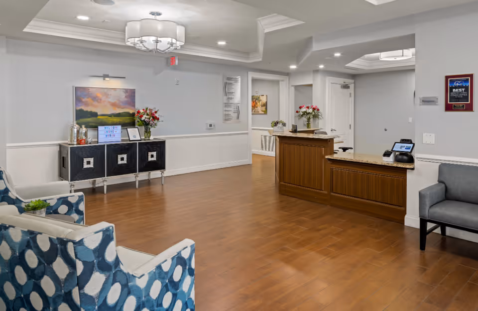 Reception area of Discovery Village At Castle Hills featuring a wooden reception desk with granite countertop, a black cabinet with decorative items and flowers, blue and white patterned chairs, a gray chair, wooden flooring, and soft lighting from ceiling fixtures.