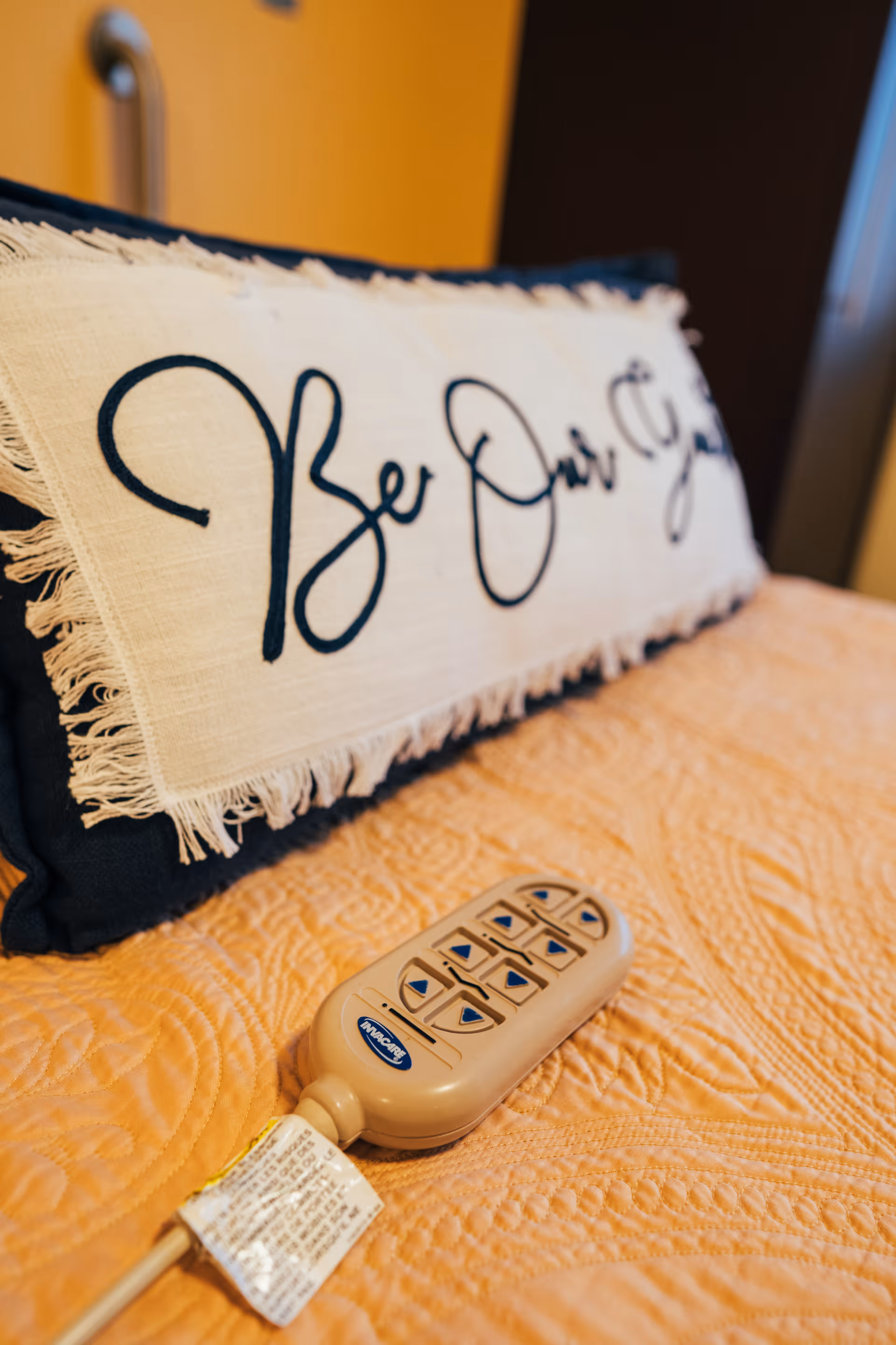 Close-up of a beige remote control with multiple buttons resting on a textured orange bedspread, with a decorative pillow in the background that has fringed edges and cursive writing.