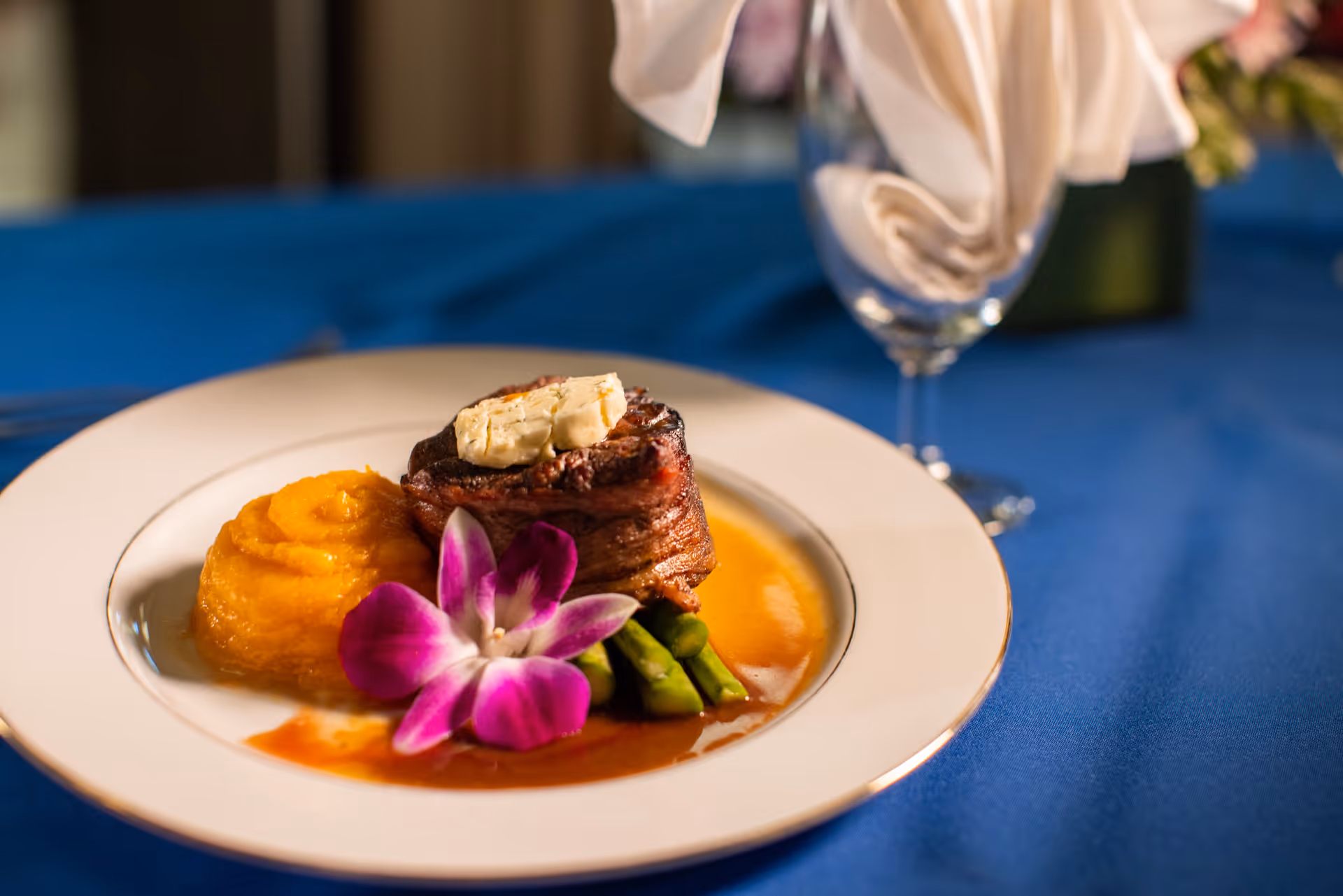 Gourmet plated steak with mashed sweet potatoes, asparagus, sauce and an orchid garnish on a white plate set on a blue tablecloth with a wine glass.