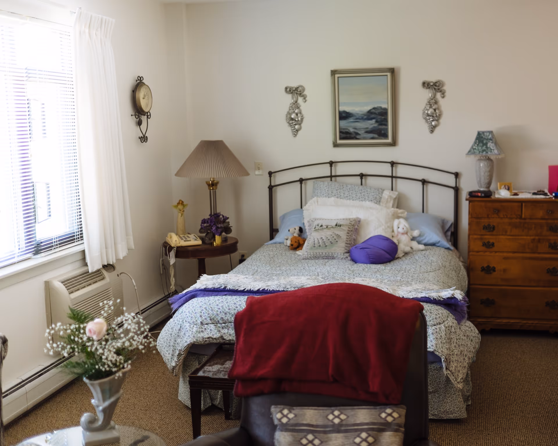 A cozy bedroom with a metal-framed bed covered in patterned bedding and several pillows, including stuffed animals. There is a wooden dresser with a lamp and various items on top, a round side table with a lamp, phone, and flowers, and a window with white curtains letting in natural light. A chair with a red blanket draped over it is in the foreground.
