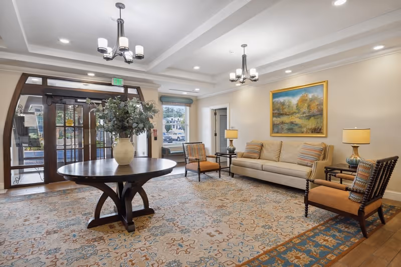 A well-lit living room area with a large patterned rug, a round wooden table with a vase of greenery in the center, a beige sofa with striped cushions, two wooden armchairs with orange cushions, two side tables with lamps, a large framed landscape painting on the wall, and glass double doors leading outside.